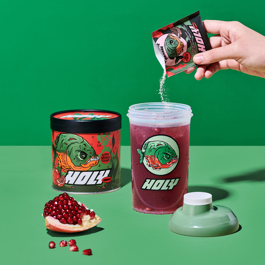 Eine Hand streut Pulver aus einer Probepackung in einen Shaker, neben einer Pappdose und einem Granatapfel.