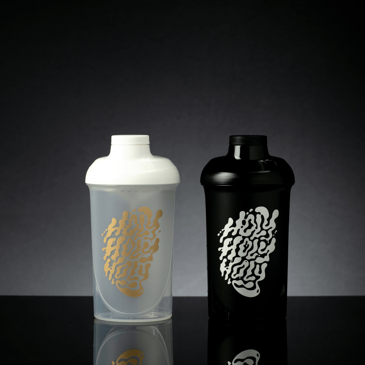 Zwei Shaker, einer weiß und transparent mit goldener Schrift und einer schwarz mit weißer Schrift, auf reflektierender Oberfläche.