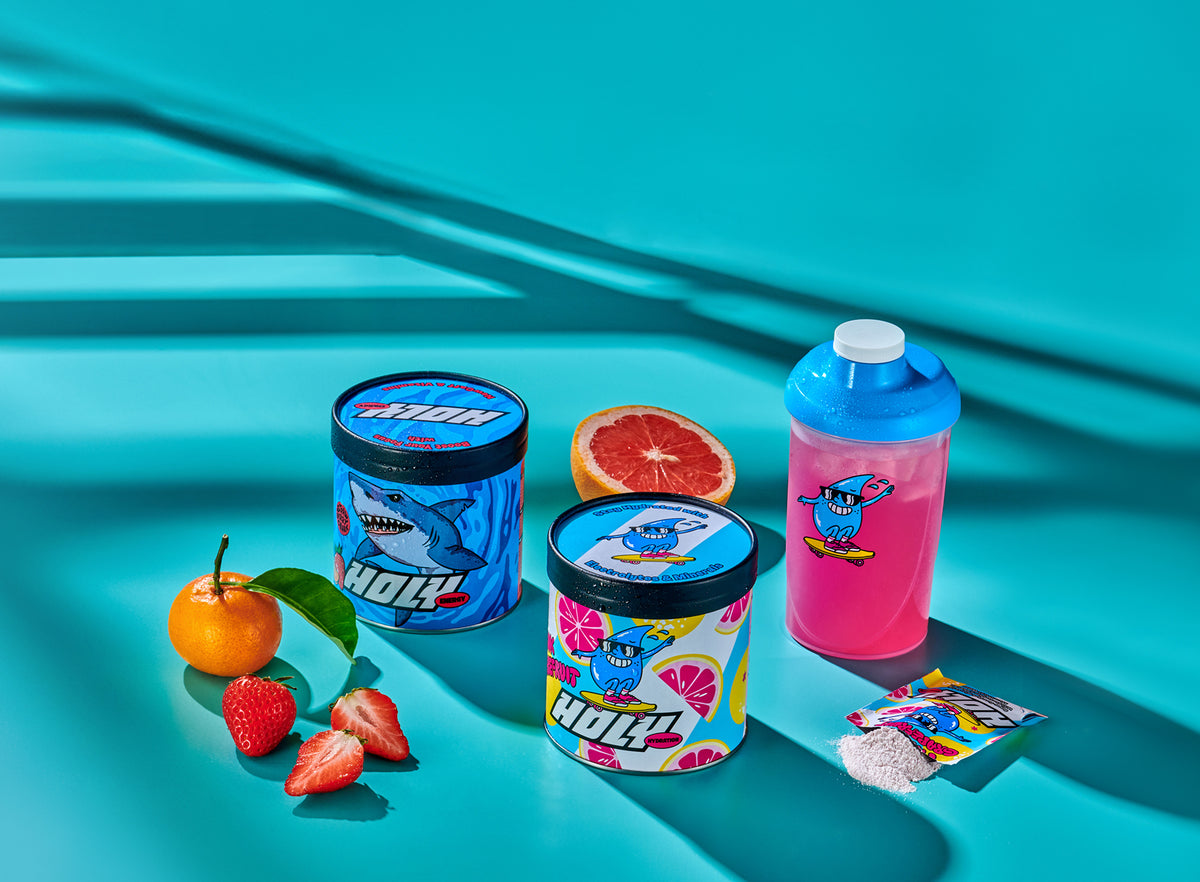 Zwei bunte Pappdosen, ein pinker Shaker, diverse Früchte und ein offenes Sachet auf türkisfarbenem Hintergrund.