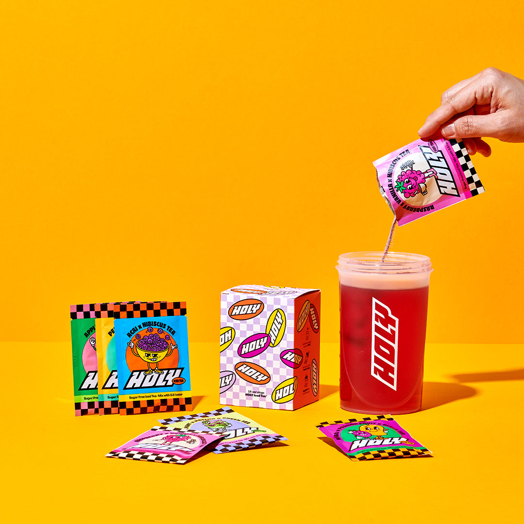 Hand gießt ein Sachet in einen Shaker, umgeben von Pappbox und weiteren Probepackungen vor gelbem Hintergrund.
