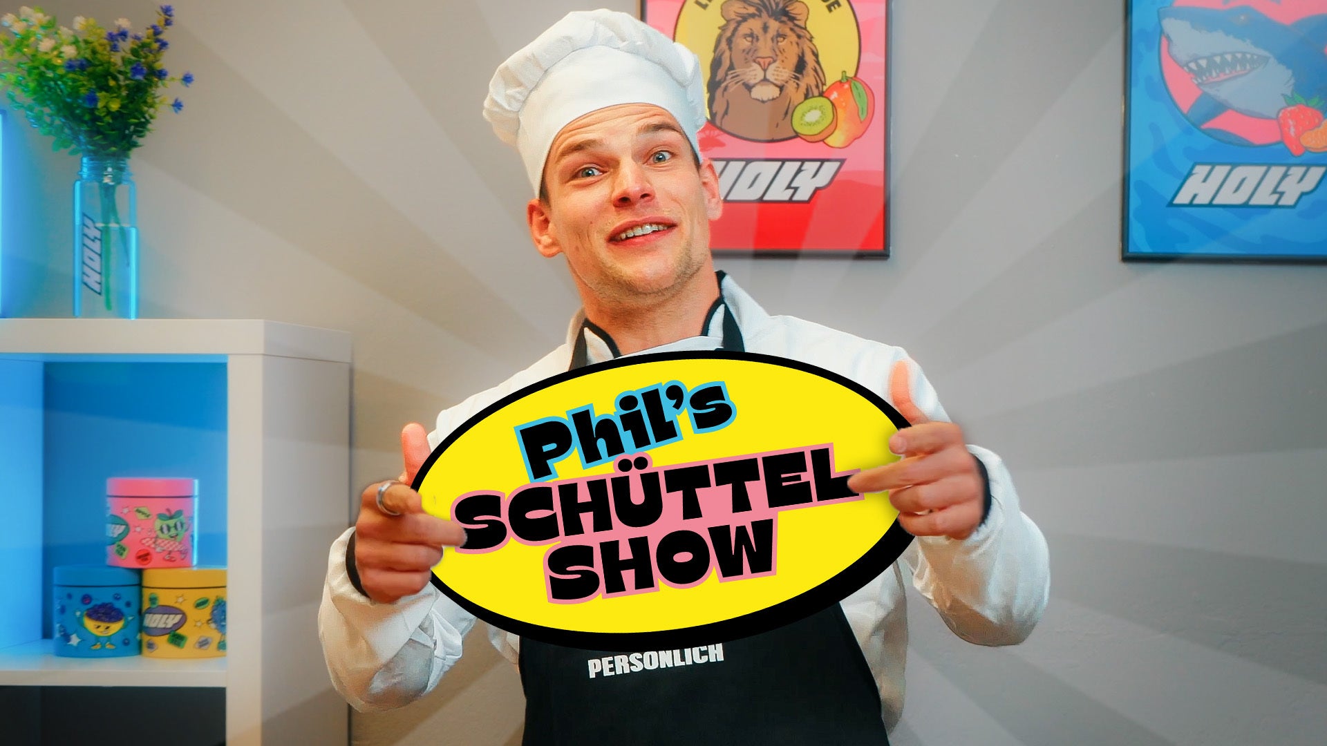 Ein Mensch in Kochkleidung hält ein Schild mit der Aufschrift "Phil's Schüttel Show" vor zwei bunten Postern.