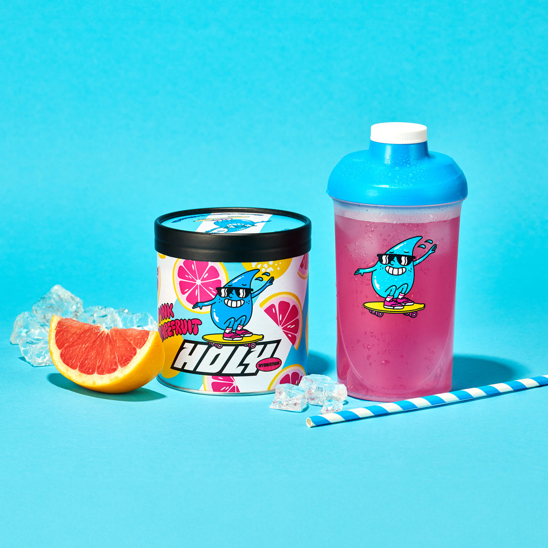 Pappdose und Shaker mit einem pinken Getränk, einem Eiswürfel, einem Strohhalm und einer Grapefruit-Scheibe auf blauem Hintergrund.