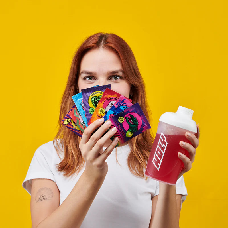 Eine Frau hält bunte Probepackungen und einen roten Shaker vor einem gelben Hintergrund.