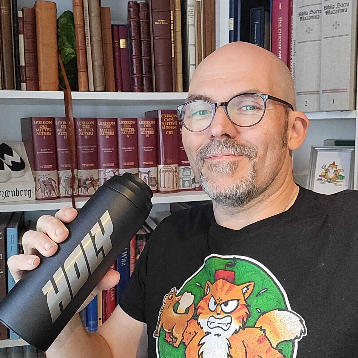 Mann in schwarzem T-Shirt hält einen schwarzen Shaker mit der Aufschrift "HOLY" vor einem Bücherregal.