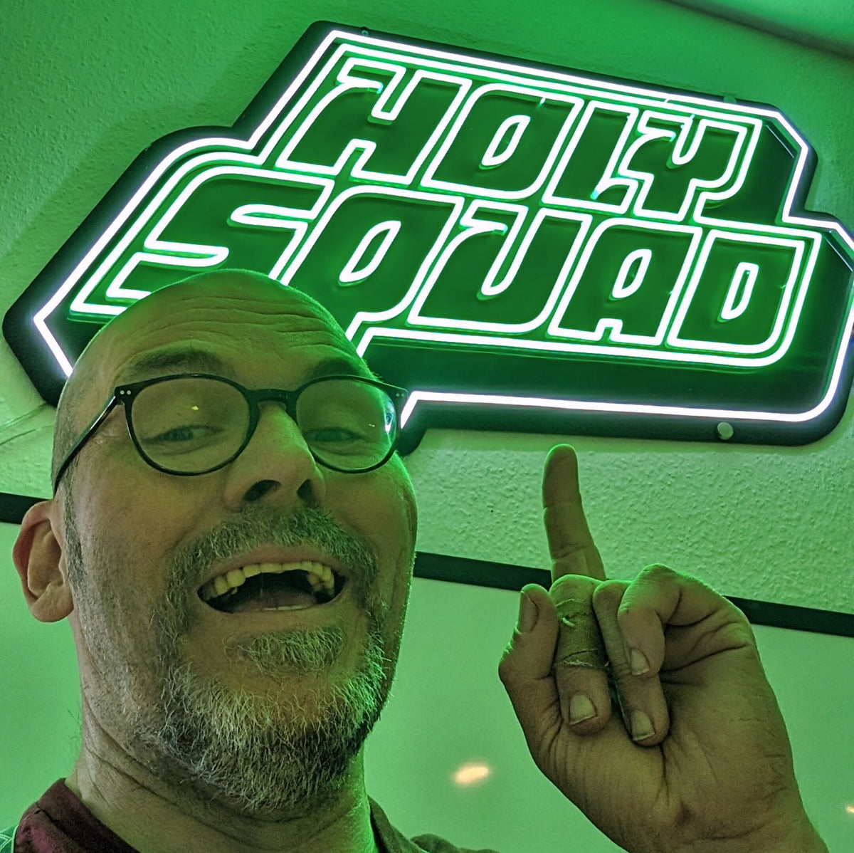 Person mit Brille zeigt lächelnd auf ein grünes Neonschild mit der Aufschrift "HOLY SQUAD".