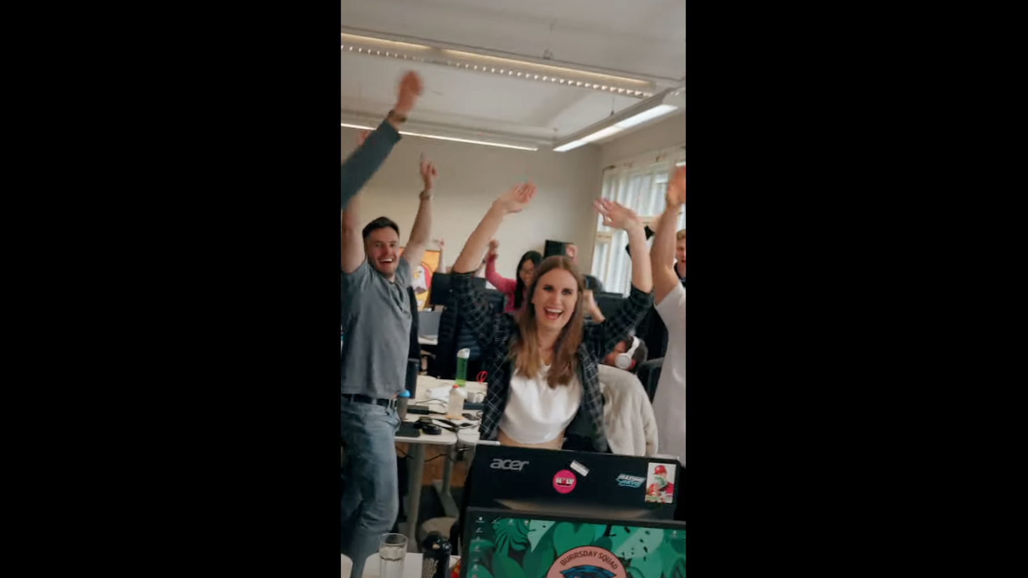 Menschen in einem Büro heben jubelnd die Arme. Im Vordergrund steht ein Laptop mit Aufklebern.