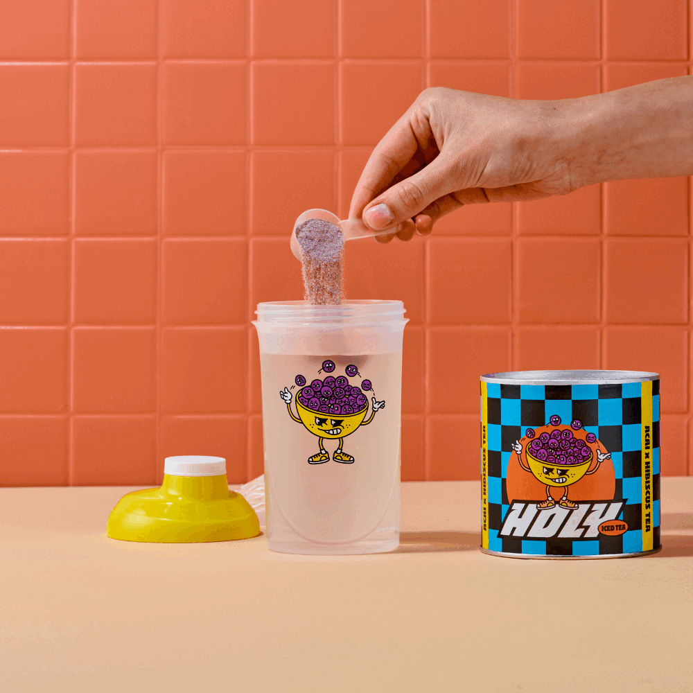 Hand fügt Pulver in einen Shaker neben einer bunten Pappdose vor einer orangefarbenen Kachelwand hinzu.
