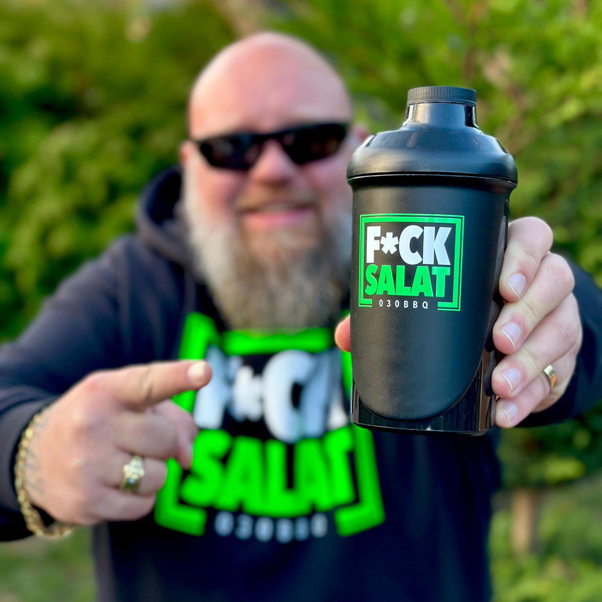 Mann mit Sonnenbrille hält einen schwarzen Shaker mit der Aufschrift "F*CK SALAT" im Freien.
